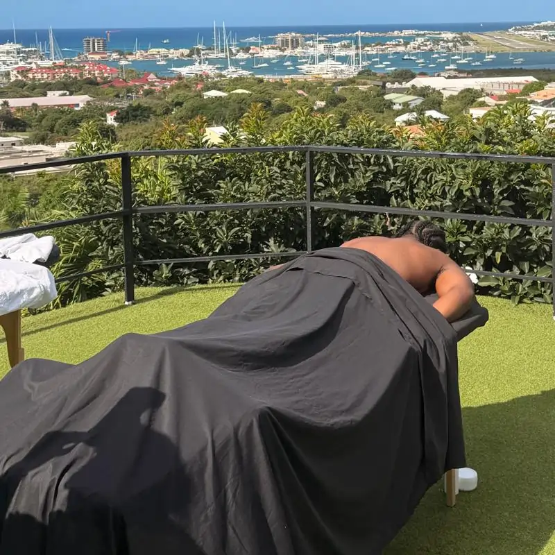 Villa Retreat A person lies face down on a massage table outdoors, covered with a dark sheet, with a scenic view of a marina, blue sea, and boats in the background. Lush greenery surrounds the area.