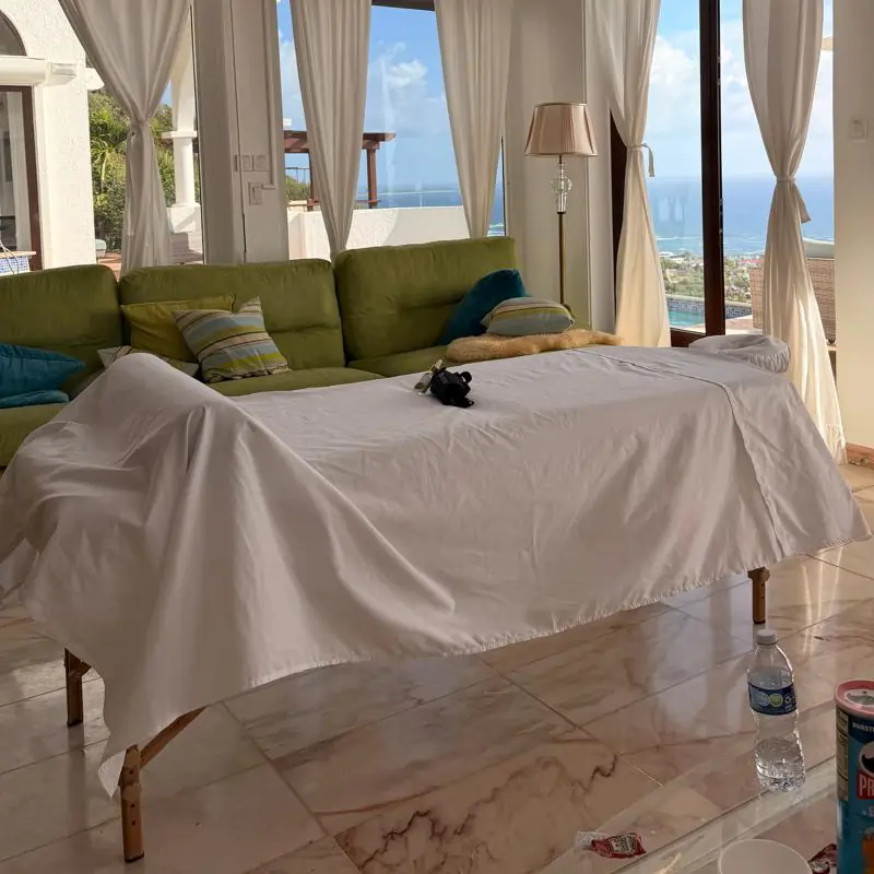 Upper Princess Villa St Maarten Massage Comfortable massage table is covered with a white sheet,in a sunlit living room with green sofas, large windows, and views of the ocean and blue sky outside.
