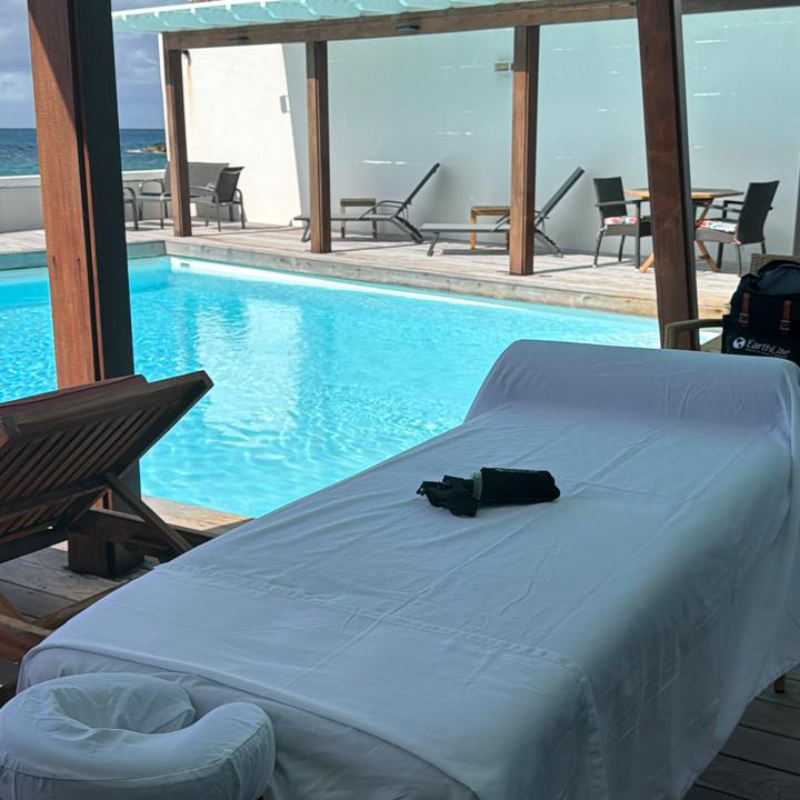 A serene poolside scene with a massage table draped in white linens, overlooking a clear blue pool. Wooden lounge chairs add a relaxing, spa-like atmosphere.