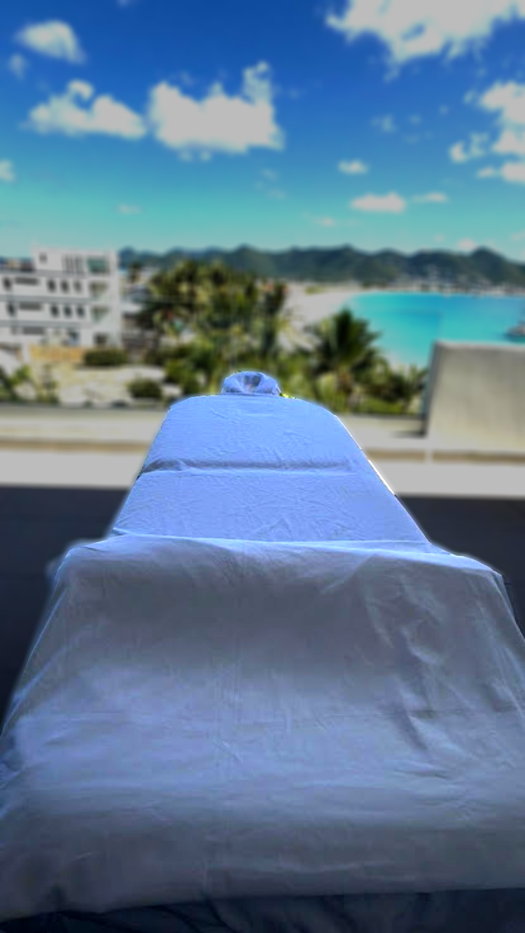 sxm massage table in beacon-hill with white sheets is set on a balcony overlooking a scenic beach view. Palm trees, blue water, and mountains are visible under a clear sky.